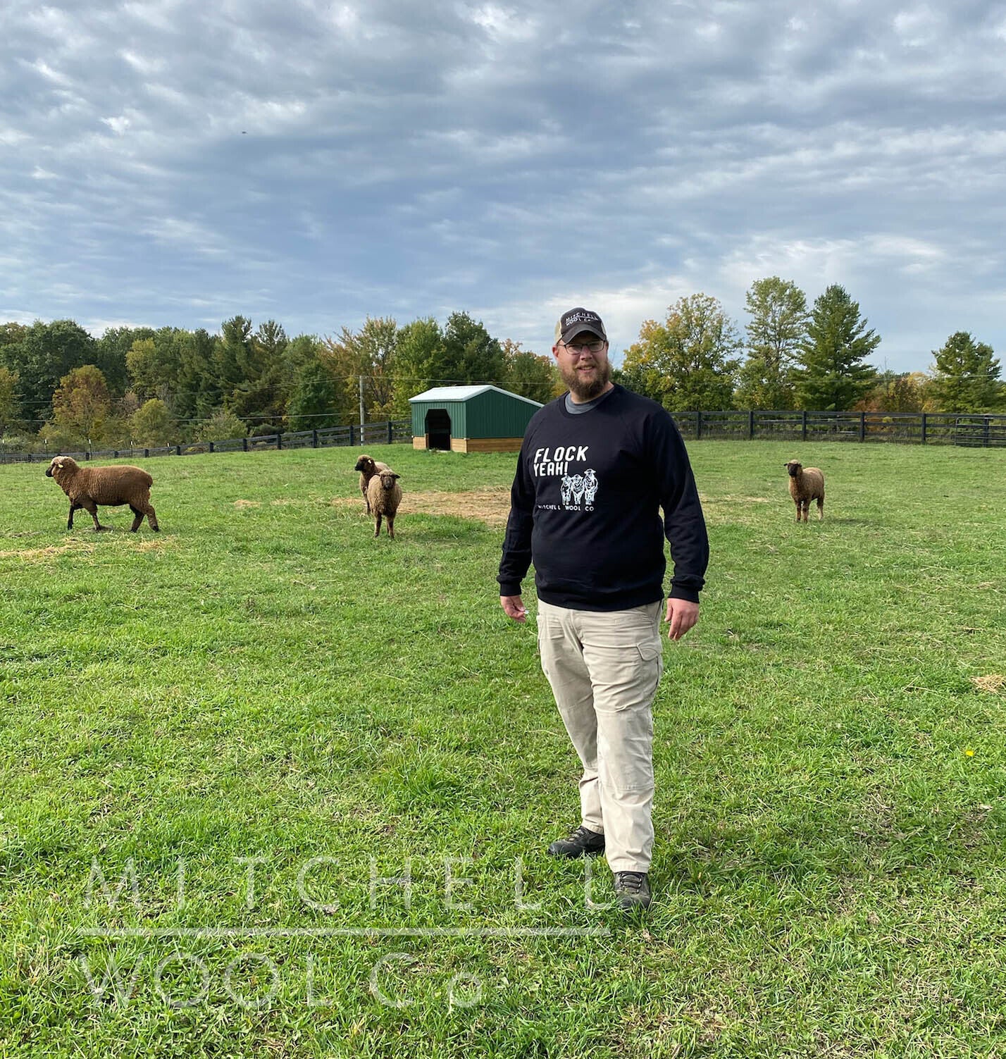 Luke stands in the pasture wearing a black sweatshirt with the words FLOCK YEAH! screenprinted in white and 3 of the Mitchell Flock sheep.