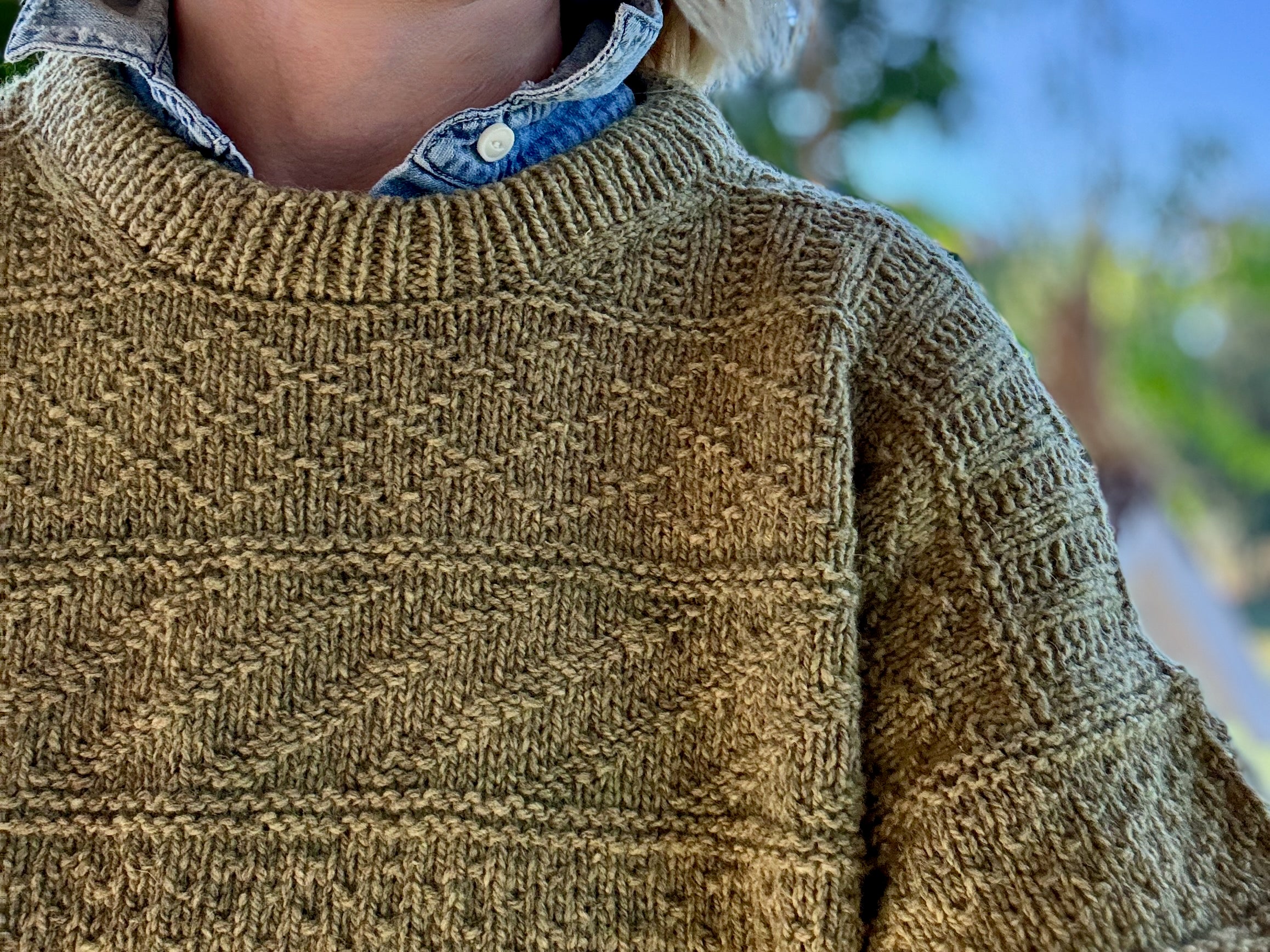 A closeup of a farm yarn knitted sweater with modern stitch patterns. It is worn by a light skinned human and 