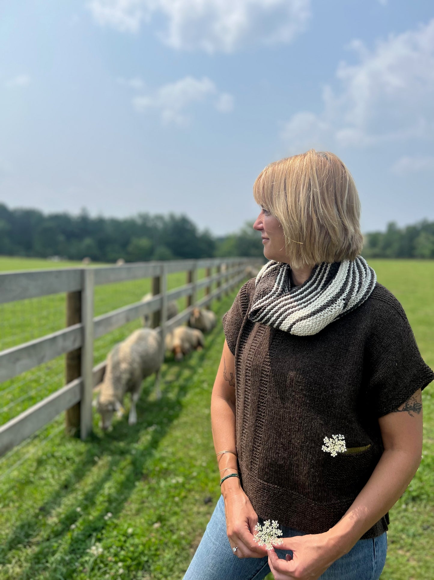 Sherry stands at the fenceline with her flock wearing a Turbulence Cowl and Alanis Sweater