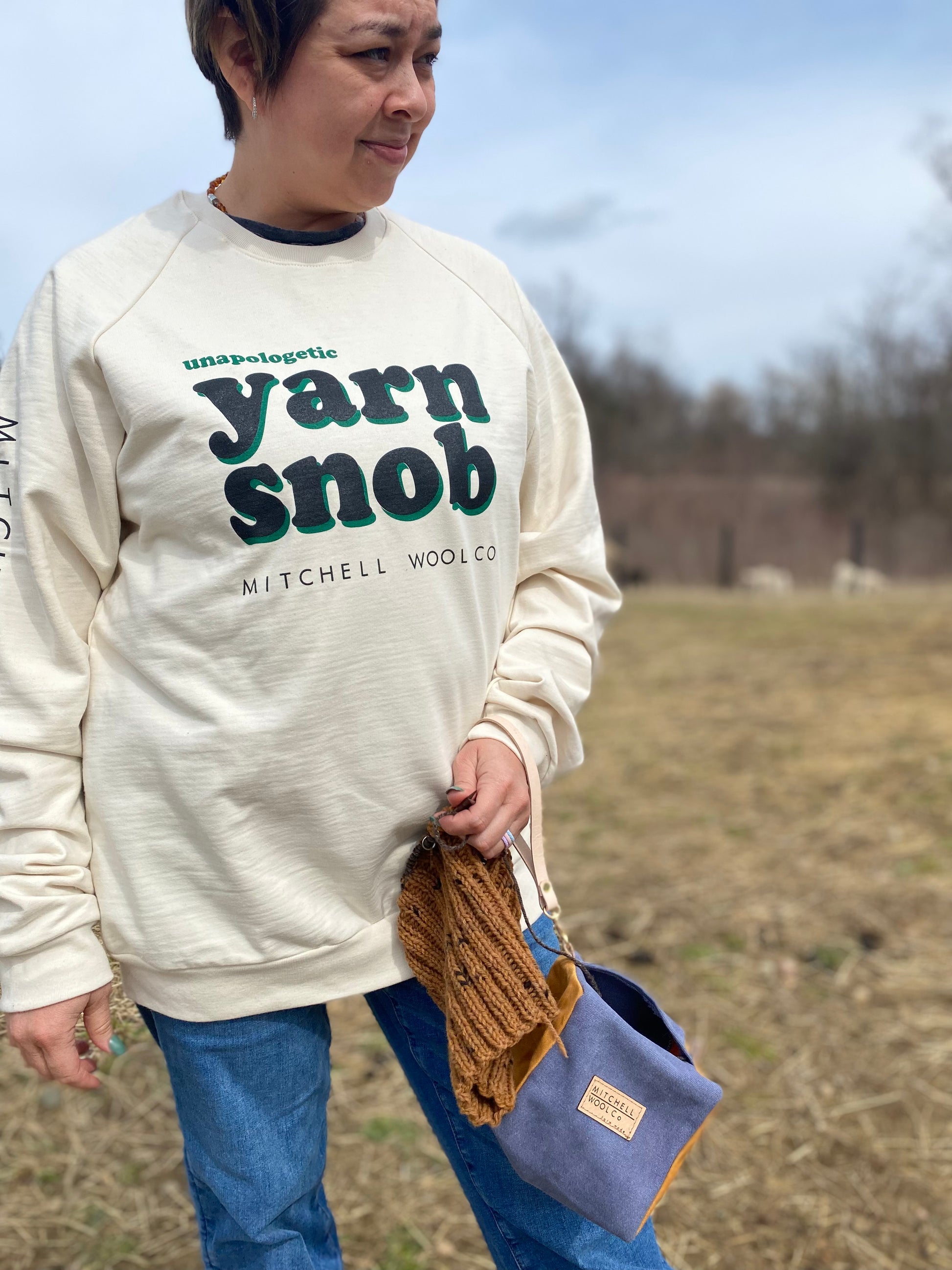 Bekah squints into the sun as she stands in the pasture wearing an organic natural colored cotton Yarn Snob sweatshirt and our yarn box knitting pouch