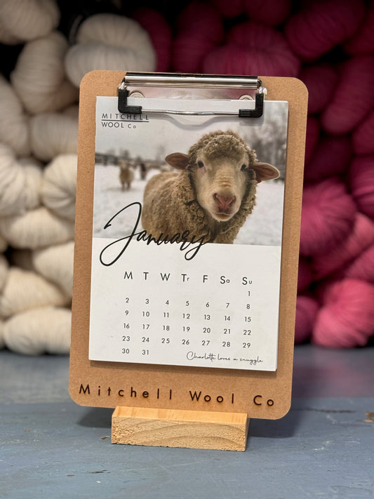 Charlotte, a happy ewe sheep stands in the snow gracing the January page of the Mitchell Wool Calendar which is a wooden clipboard on a pine block stand. It sits in front of stacked skeins of pink and white yarn on a blue cabinet.
