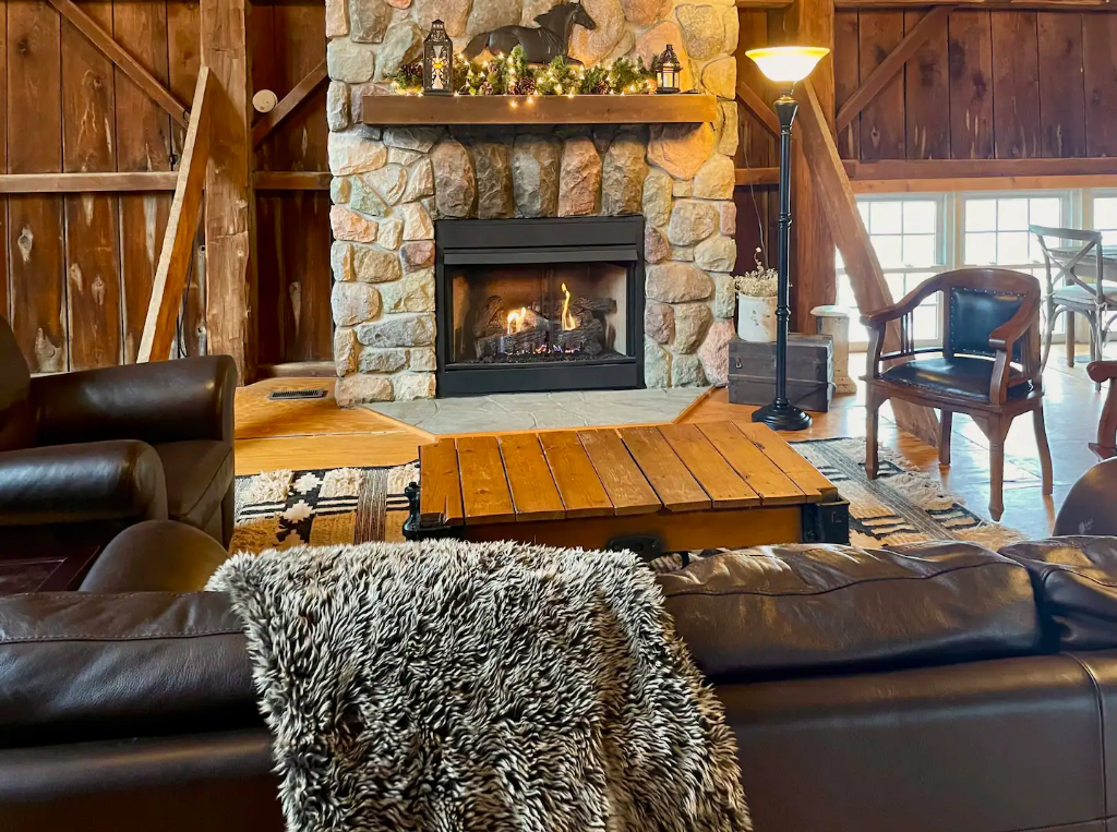 Won't this be so fun to meet new friends around the fire? A stone fireplace centers the room of a cozy light filled barn living room with leather sofas and sheep skin pelts