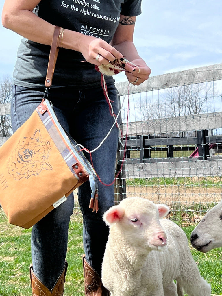 Sherry stands knitting in the field with a Betty project bag on her forearm and knitting a sock from wool she grew herself. Well her sheep did, but we raised them so we're mighty proud.