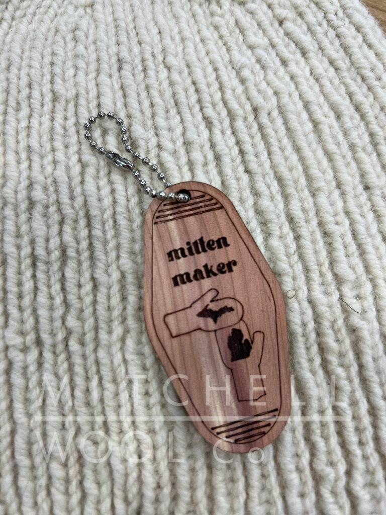 I'd knit thatThe words Mitten Maker are above the shape of Michigan etched onto 2 mittened hands sketched on a wood vintage style hotel key chain sits on a hand knit cormo natural hat.