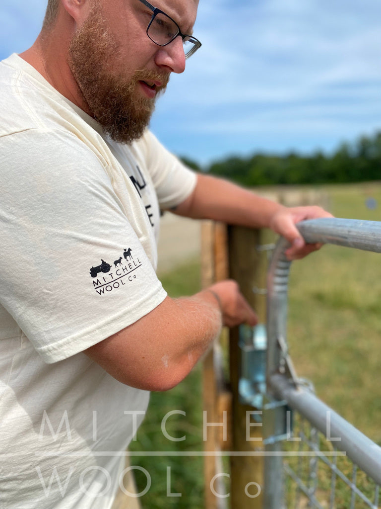 Luke opens the gate to rotate our flock into a fresh new pasture. He is wearing an organic natural colored cotton tee shirt that shows the Mitchell Wool Logo and a farmer on tractor, horse, chicken & sheep on the sleeve in black ink. The shirt was made in the USA 