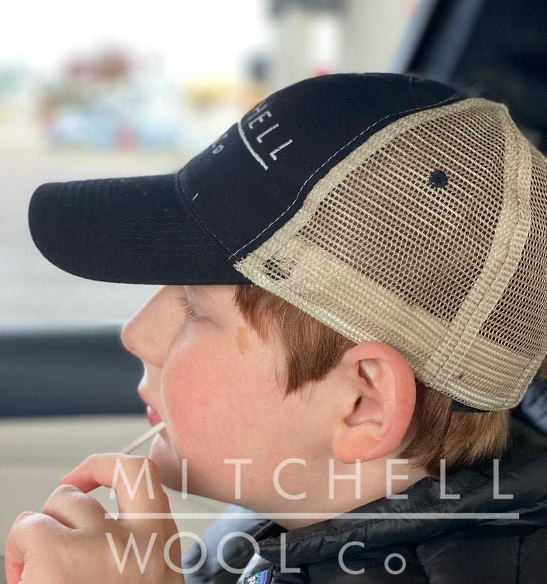 Side view of Future Farmer wearing his MWC black and natural trucker hat. He sucks on a lollipop and his adorable red locks pop out the bottom of the mesh.