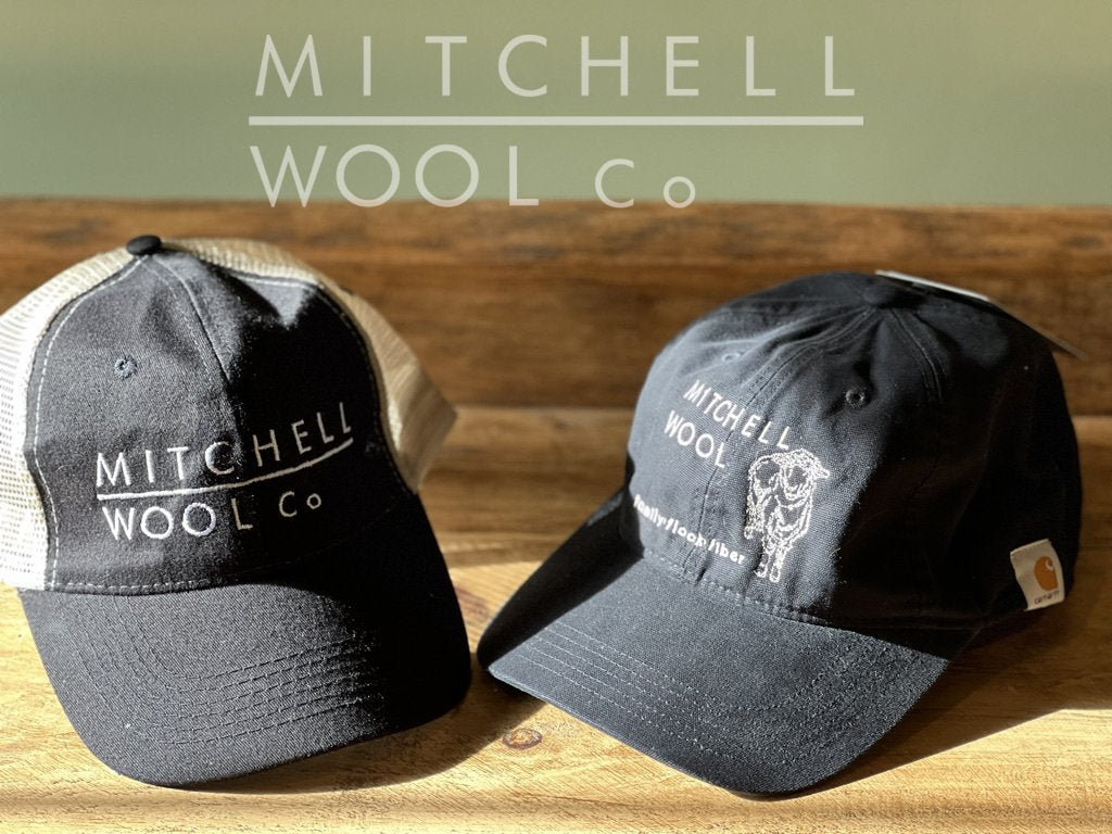 The MWC black & natural trucker hat sits to the left of our Black cotton Carhartt hat on a beautiful old pine table with warm sunshine shining upon them.