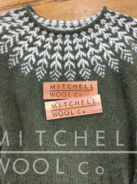 Two cedar planks engraved with the Mitchell Wool Co logo sit on a colorwork sweater on an old pine table. The sweater is green with white feather details
