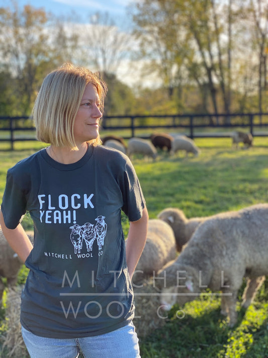 Sherry looks off across the pasture wearing a Flock Yeah! organic charcoal hemp/viscose tee shirt. Her flock of sheep happily graze the pasture behind.