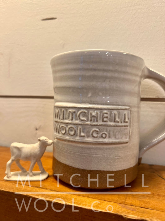 A hand thrown white glazed Mitchell Wool Co. coffee mug sits on a wood shelf in front of a white shiplap wall with a tiny lamb figurine next to it. The glaze beautifully highlights the soft rings around the body and compliments the raw pottery bottom. The inside is fully glazed.