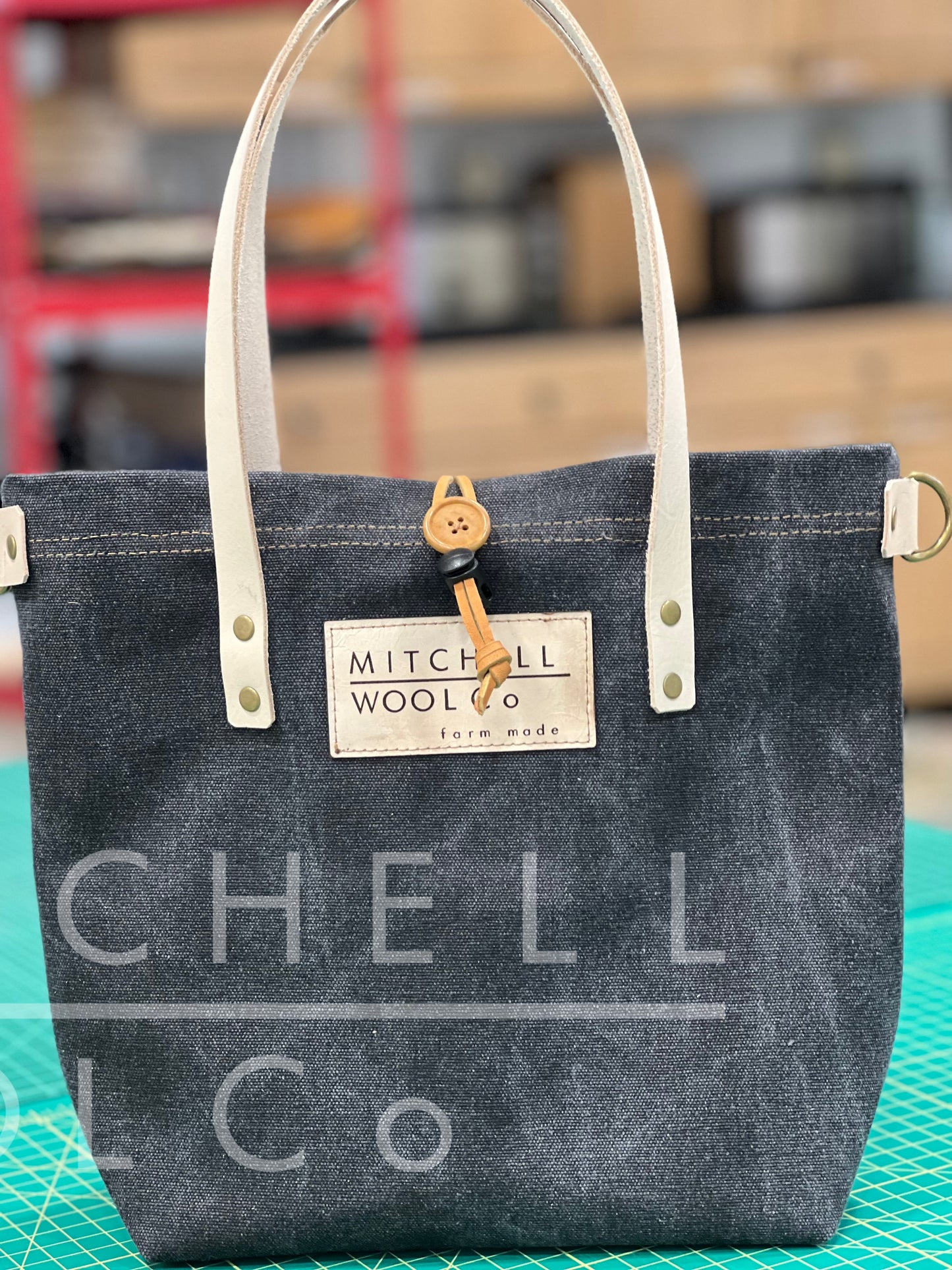 a Simple Tote made from black washed canvas denim with undyed vegetable tanned leather handles sits on a work table. it has D rings at the side for carrying with a crossbody strap and a MWC patch in leather on the front. The bag is closed with a leather strap and button toggle.