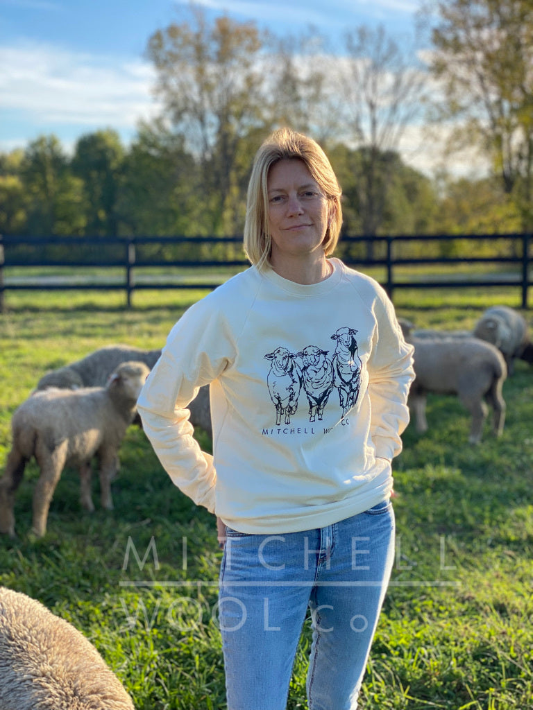 Sherry stands amongst her flock on a sunfilled afternoon wearing a natural cotton sweatshirt bearing the Mitchell Wool Co logo and a sketched screen print of 3 cormos in black ink.