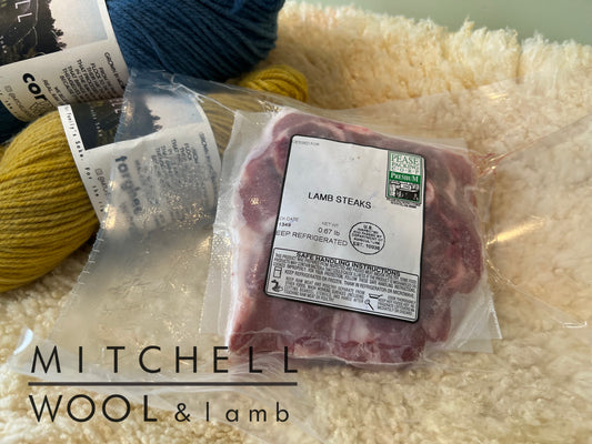 a richly marbled pasture raised lamb steak package sits atop a soft natural white wooly lamb fleece. behind it is a skein of marigold cormo yarn