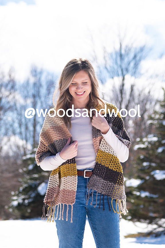 Melissa, a white woman with blond hair stands modeling her Interwoven Scarf that she designed for crochet. It is in the colors of raw brown, Farm Friends, Marigold, Natural Raw and Avocado plant dyed yarns